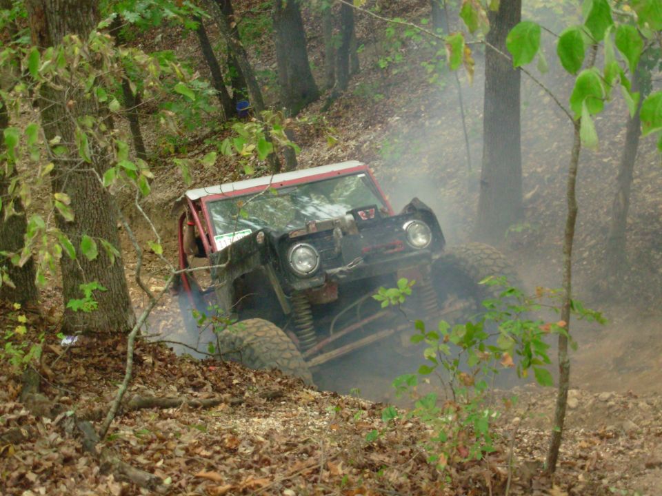 Red fenderless trail rig climbing dusty wooded hill on oversized mud tires