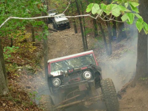 1983 Ford Bronco - Multiple off-road vehicles navigate a challenging dirt trail through a forest, kicking up dust.
