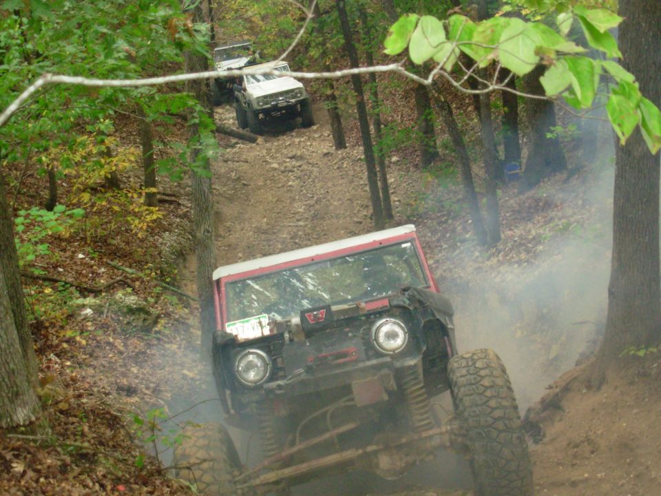 Red and white off-road truck flexing up a dusty rocky hill climb in the woods