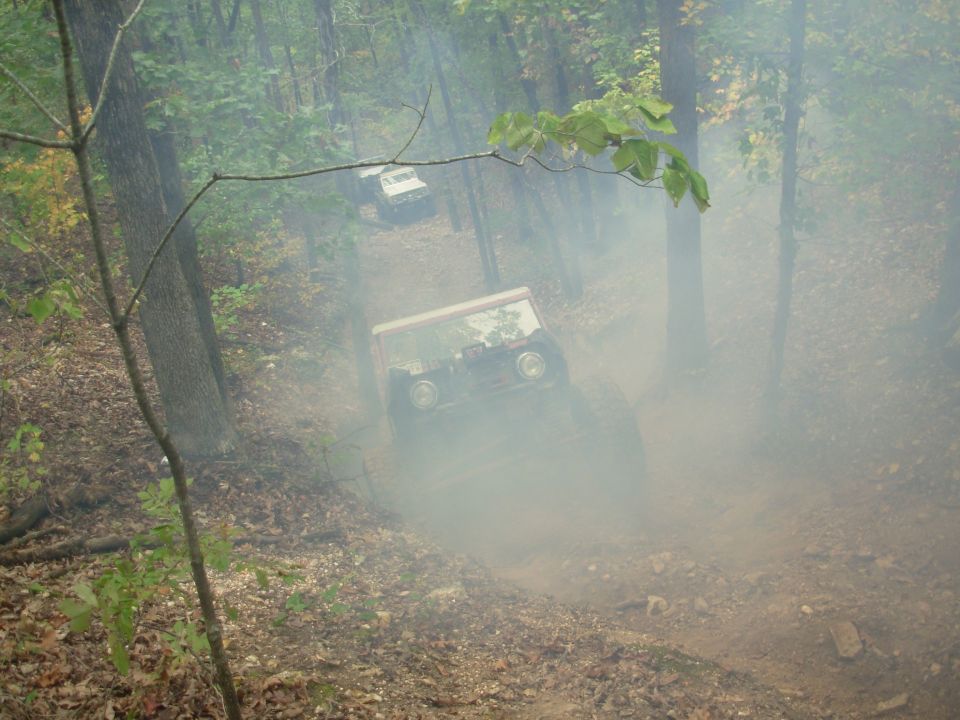 White and red caged 4x4 climbing a dusty wooded trail with a second rig waiting uphill