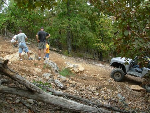 1983 Ford Bronco - Silver off-road SUV navigates a challenging rocky terrain with trees in the background.
