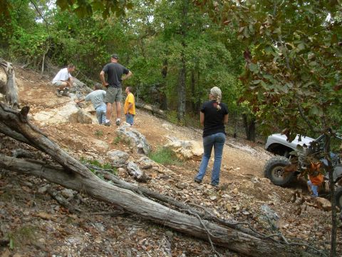1983 Ford Bronco - People observe off-road vehicles tackling a challenging, wooded, rocky incline.