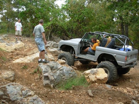 1983 Ford Bronco - A heavily modified Ford Bronco is shown off-roading on a rocky trail with two people observing.