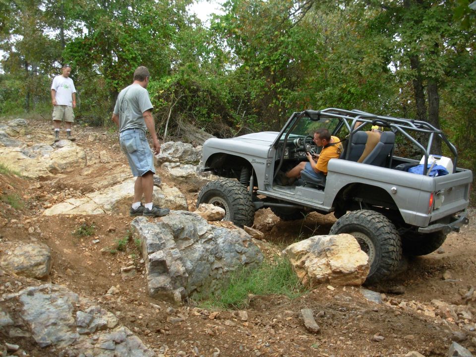 Gray caged topless early Ford Bronco on rock ledge with spotters watching