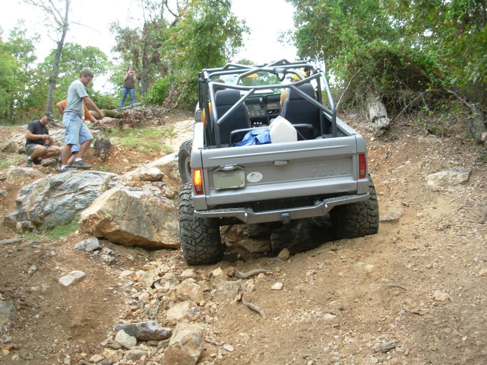 Silver early Ford Bronco with roll cage climbing rocky trail ledge, rear view, spectators nearby