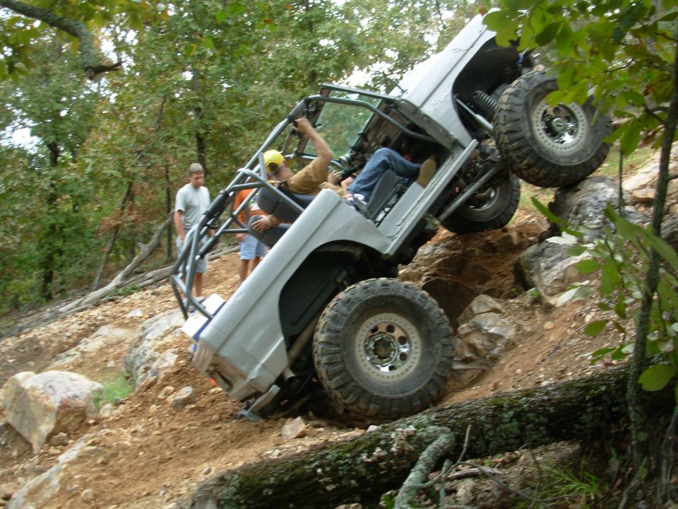 White caged open-top 4x4 on mud-terrain tires climbing a steep rock ledge with spotters watching