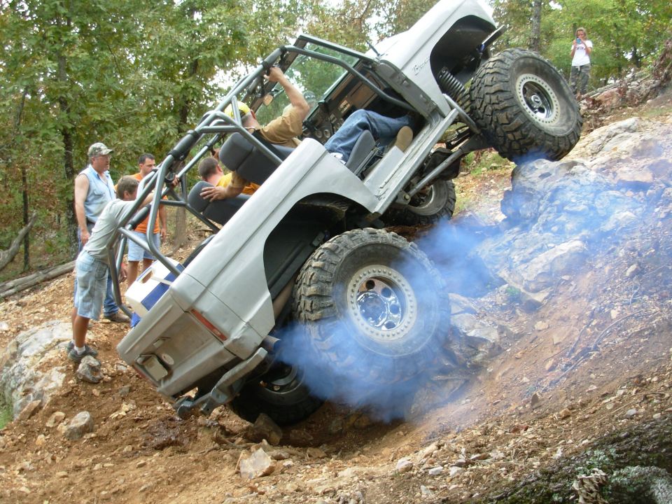 White caged Bronco climbing a steep rock face with tires smoking, spectators watching