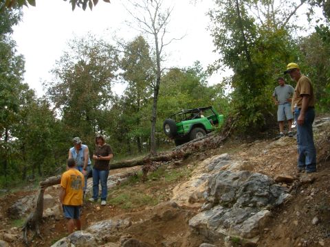 1983 Ford Bronco - Off-roading enthusiasts watch a green Ford Bronco tackle a challenging, wooded terrain.