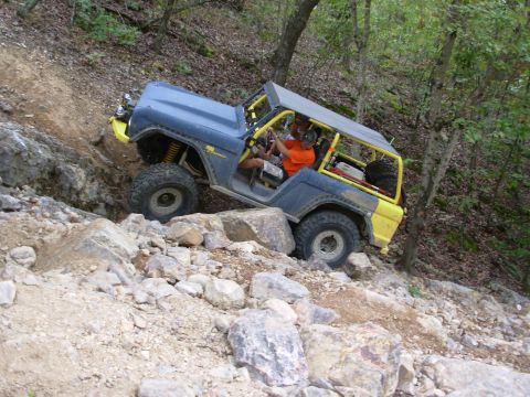 1983 Ford Bronco - An off-road SUV navigates a rocky incline with a yellow roll cage and spare tire.