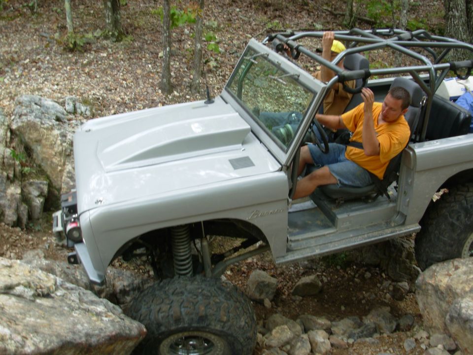 Gray topless early Ford Bronco with roll cage and coilovers flexing over trail rocks