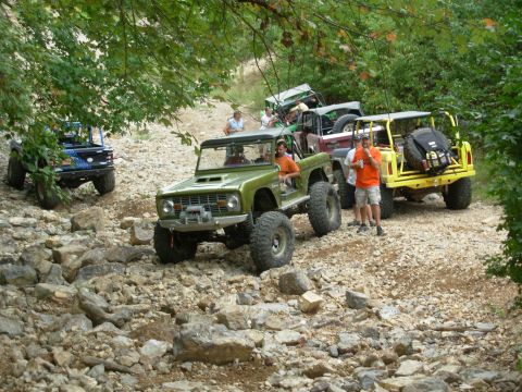1983 Ford Bronco - Several modified off-road vehicles are seen navigating a challenging, rocky trail surrounded by trees.