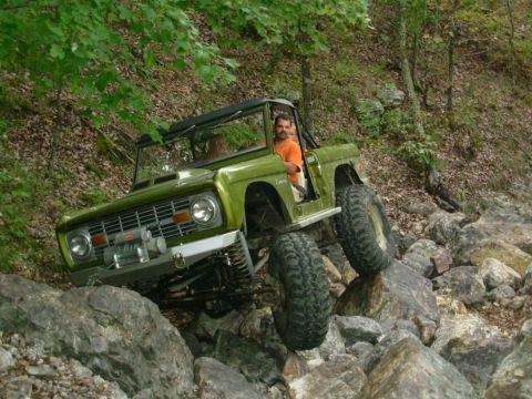 1983 Ford Bronco - Off-road vehicle tackling a difficult rocky path with trees and foliage in the background.