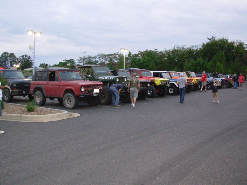 Red early Ford Bronco leading a row of lifted Jeeps at an evening 4x4 meet