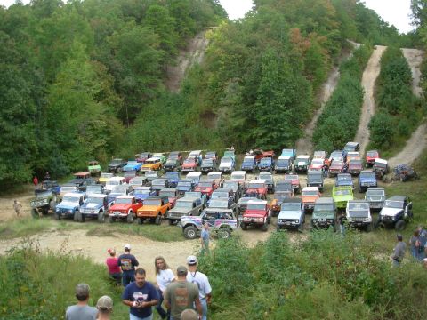 1983 Ford Bronco - A large group of off-road vehicles gathered in a natural setting with wooded hills.