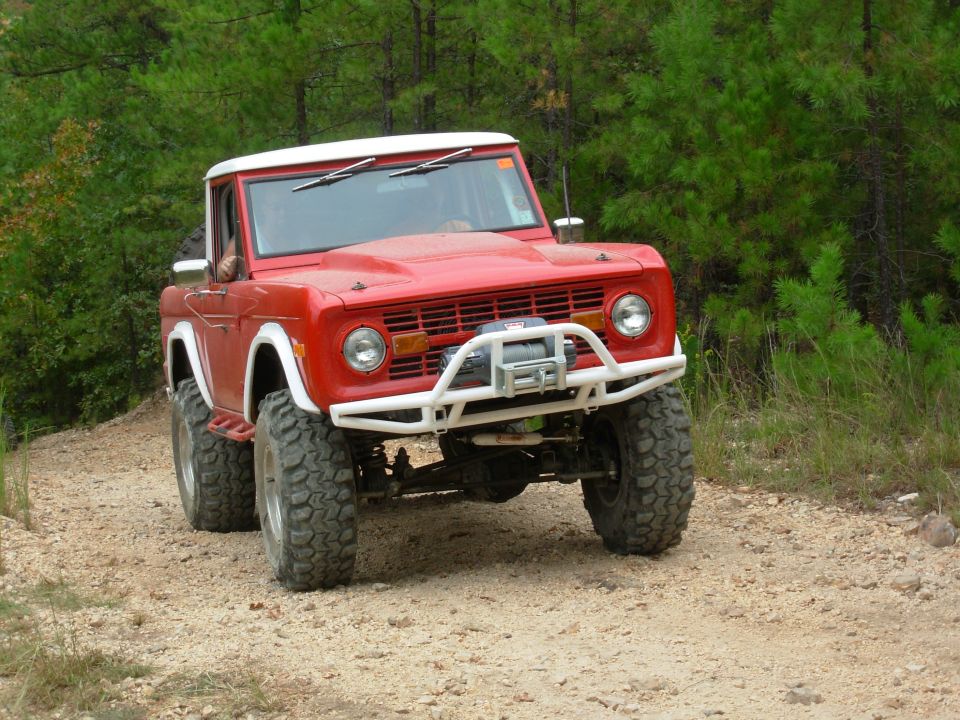 Red early Ford Bronco with white roof and white tube bumper on a gravel forest trail