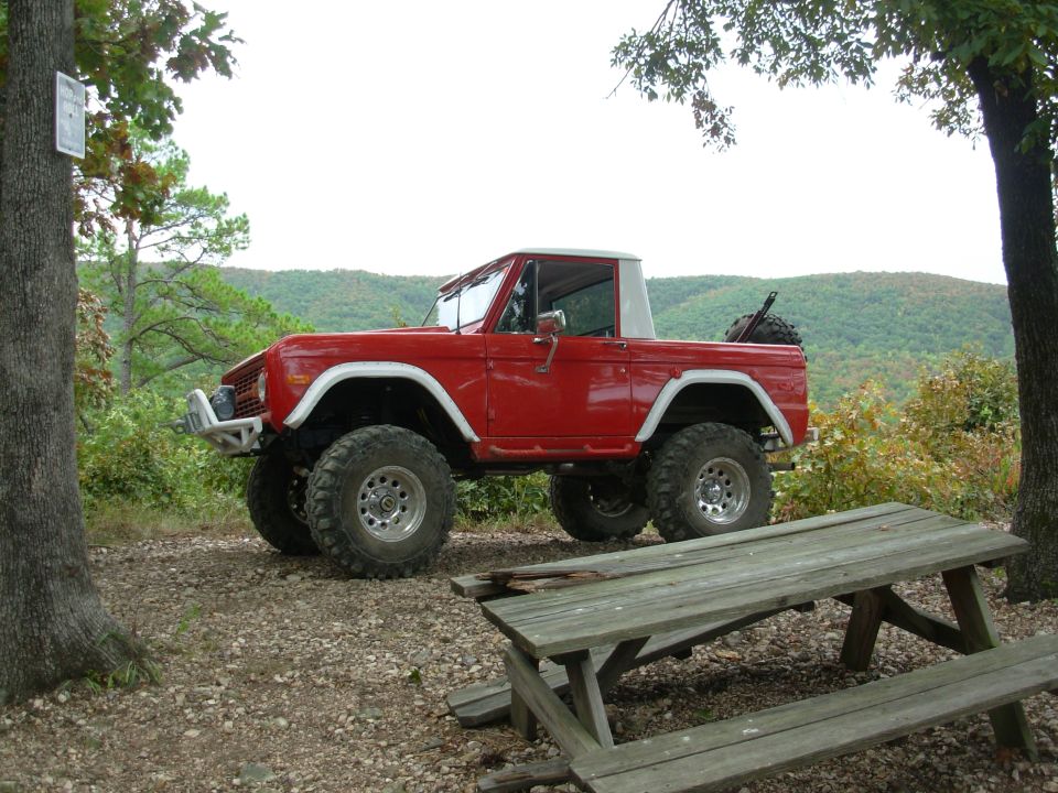 Lifted red early Ford Bronco half-cab with white roof at a mountain gravel overlook