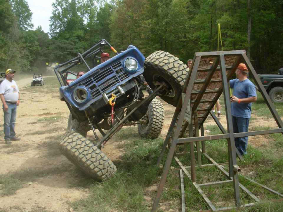 Blue caged early Bronco climbing a steel ramp obstacle at an off-road event