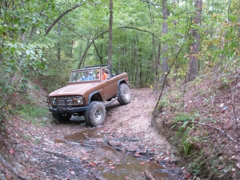 1983 Ford Bronco - A brown Ford Bronco navigates a wooded trail with a stream crossing.