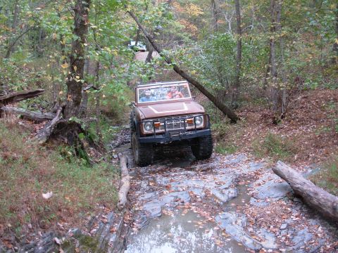 1983 Ford Bronco - A Ford Bronco navigates a challenging off-road trail with fallen logs and water obstacles.