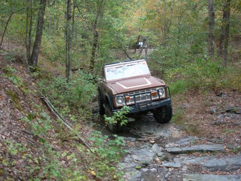 1983 Ford Bronco - A brown Ford Bronco navigates a challenging rocky path through a dense forest.