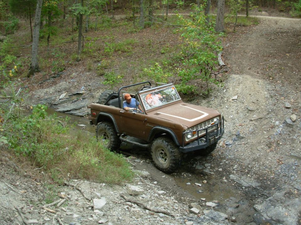 Brown topless early Ford Bronco crossing a muddy creek on a wooded trail