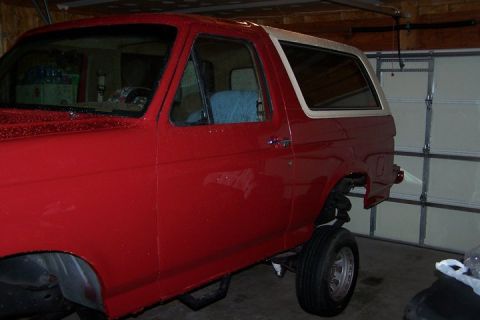 1990 Ford Bronco - This red Ford Bronco is parked in a garage, showing its side profile with a lifted suspension and missing rear components.