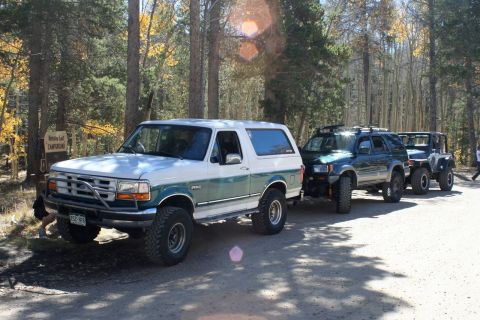 1996 Ford Bronco - A white and green 1996 Ford Bronco leads a line of SUVs on a dirt road near a campground.
