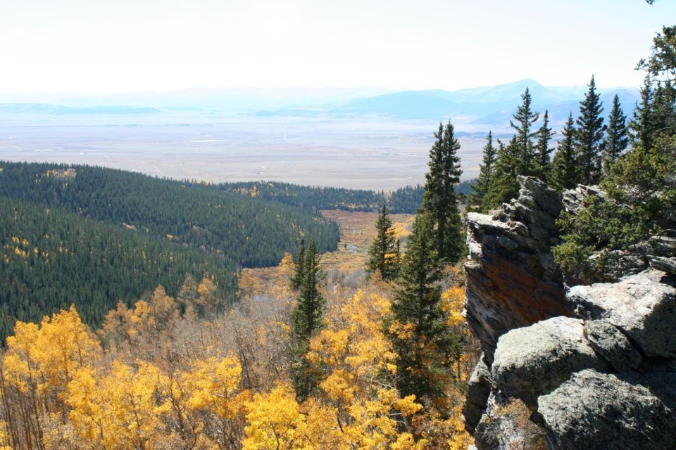 Mountain landscape with yellow and green trees, rocky cliff, and distant plain under a hazy sky