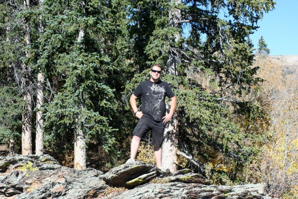 Man in t-shirt and shorts standing on rocks with pine trees behind him