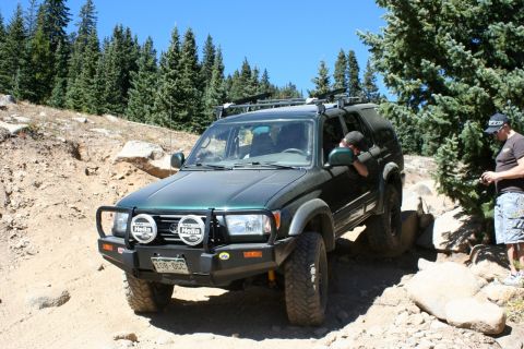 1996 Ford Bronco - A green SUV navigates a challenging rocky terrain on a sunny day with trees in the background.