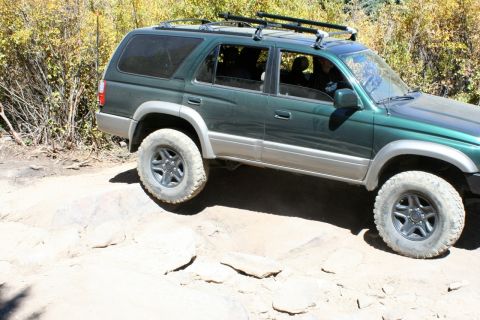 1996 Ford Bronco - Green SUV navigates a rocky trail with its front passenger side wheel elevated.