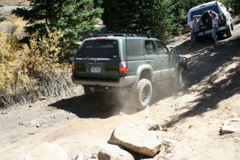 1996 Ford Bronco - Two SUVs navigate a challenging off-road trail, kicking up dust on a sunny day.