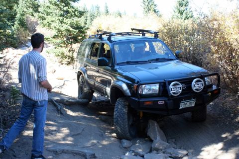 1996 Ford Bronco - A man watches as a dark SUV navigates a challenging, rocky off-road path.