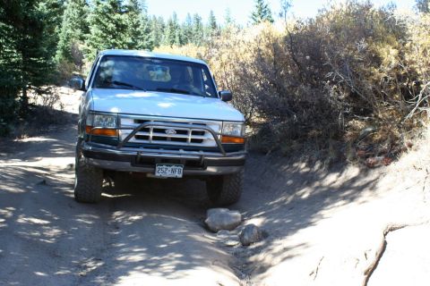 1996 Ford Bronco - A white Ford Bronco navigates a rocky dirt path surrounded by trees and brush.