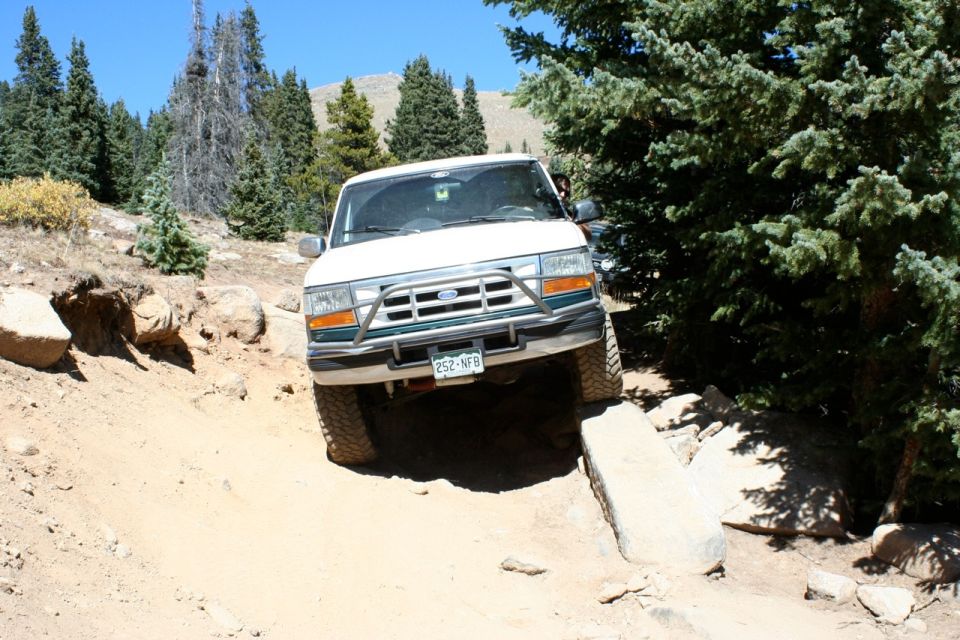 White 1992 Ford Bronco driving on a rocky dirt trail with trees and mountains in the background