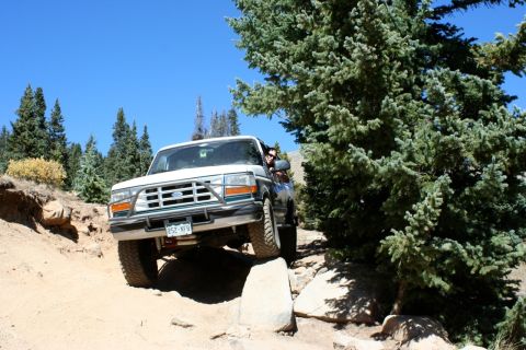 1996 Ford Bronco - A white 1996 Ford Bronco tackles a challenging off-road path with large rocks and trees.