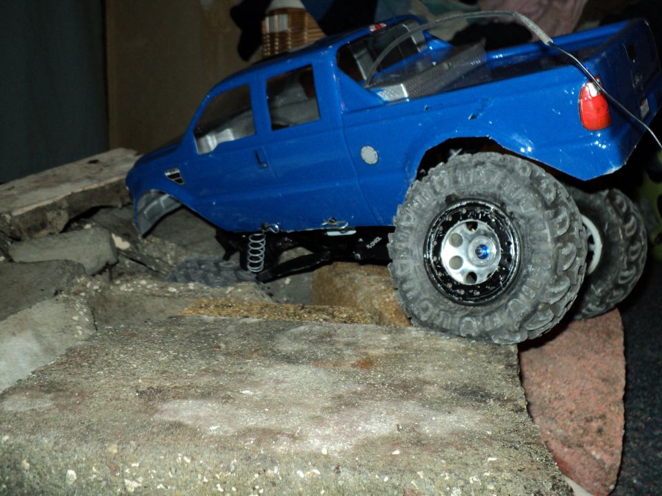 Blue RC crawler pickup climbing broken concrete, showing links and crawler tires