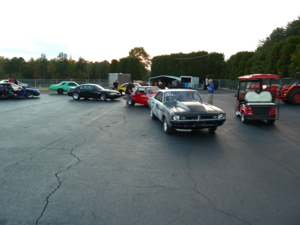 Several cars are parked on asphalt, including a black car, a red car, and a drag car with a hood scoop. A red golf cart and a tractor are also visible