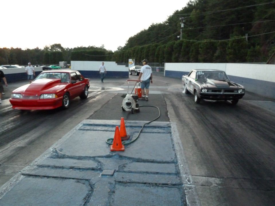 Red drag car with a large hood scoop on a race track, viewed from the front-right