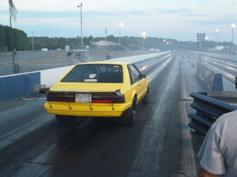 1992 Ford Mustang - A yellow drag car sits on the starting line of a drag strip under a cloudy sky.