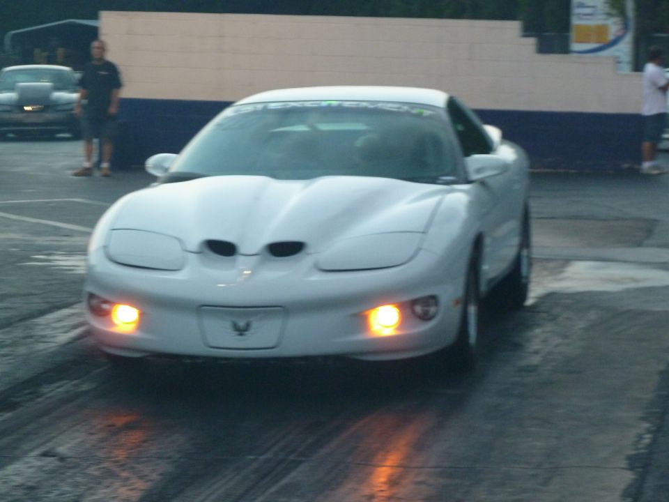White Pontiac Firebird Trans Am on drag strip. Windshield banner reads 'GET EXCITEMENT'. Another car is visible in the background