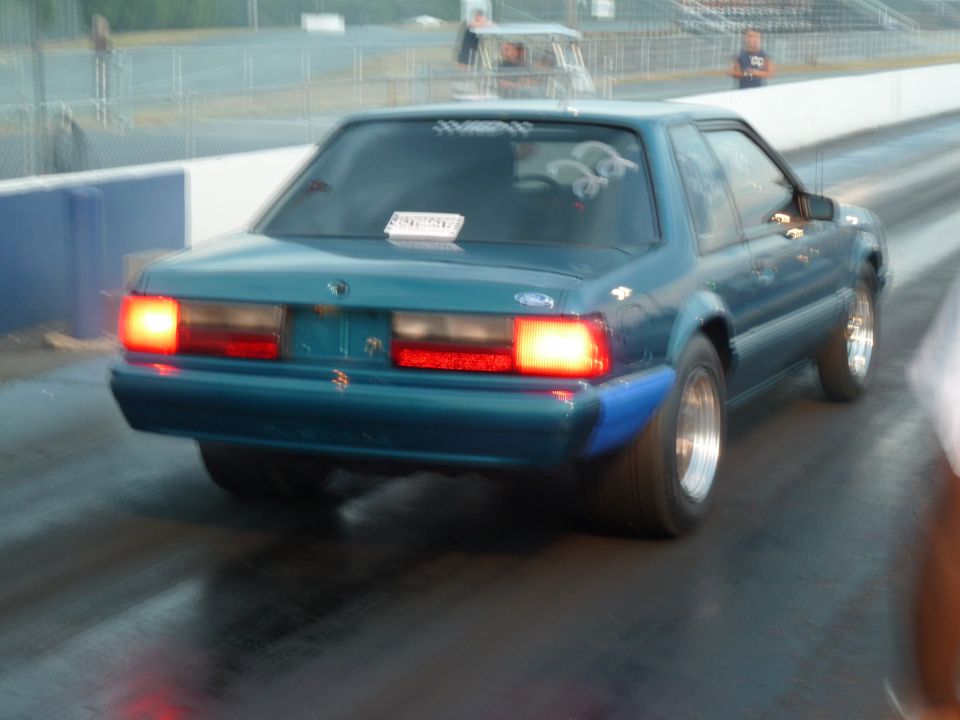 Teal coupe with blue fender flares and racing slicks on a drag strip, rear view