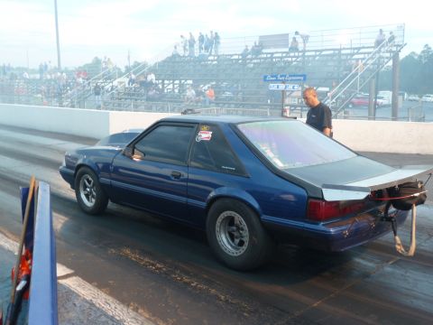 1992 Ford Mustang - A blue 1992 Ford Mustang coupe is staged at a drag strip, ready for a race, with spectators in the background.