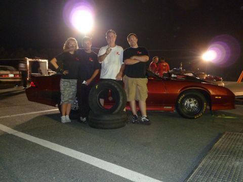 1992 Ford Mustang - A group poses with a red drag car and spare tires at a nighttime event.