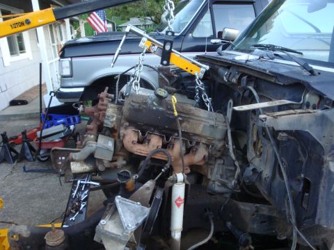 1989 Ford Bronco - A grimy V8 engine hanging from a half-ton engine hoist and leveler bar, pulled out of a truck whose stripped engine bay, bare firewall and…