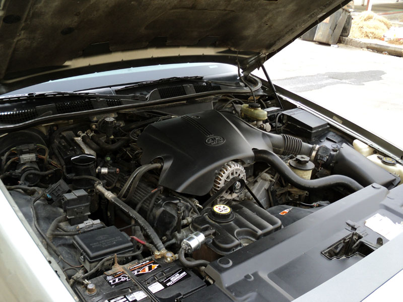 Engine bay of a light gold sedan showing a 4.6L V8 with black engine cover, alternator, and battery