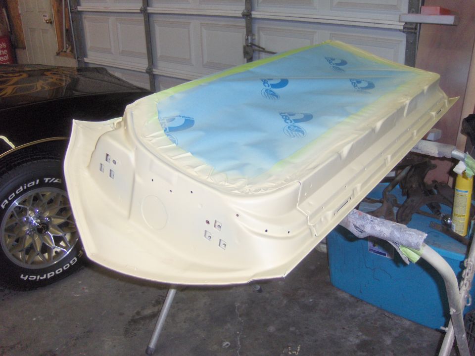 Car door panel covered in blue plastic and taped off, sitting on a stand in a garage. A black car tire is visible to the left