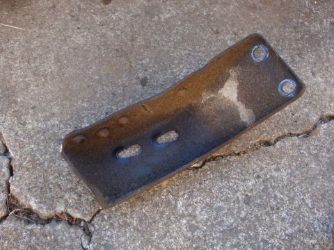 1989 Ford Bronco - A stamped steel bracket laid on cracked concrete, its rusty-brown surface still showing traces of black paint. The plate has two round bolt…