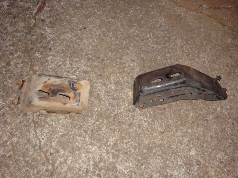 1989 Ford Bronco - Two stamped-steel engine mount brackets lying side by side on a concrete driveway, both drilled with slotted and round bolt holes. The left…