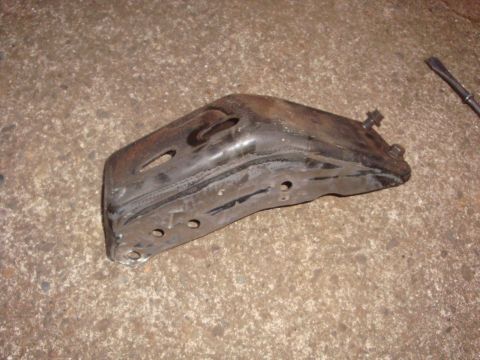 1989 Ford Bronco - A greasy stamped-steel frame bracket lying on a concrete garage floor, showing a welded seam along its length, a slotted adjustment hole, s…